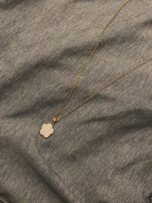 Guldigt halsband med blomma - Snyggt halsband i guld med tunt kedja och blomformad berlock. Ena sidan av berlocken är slät och ljus, medan andra sidan har ett detaljerat blommönster. Perfekt accessoar för att lyfta din outfit.