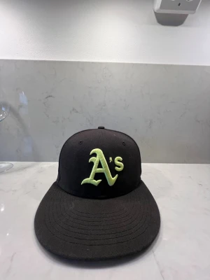 Svart New Era Oakland A's keps - Säljer en svart keps från New Era med broderad limegrön Oakland A's-logga framtill och New Era-logga på sidan. Klassisk rak skärm och justerbar passform. Perfekt för dig som gillar sportig streetstyle och vill sticka ut med en ikonisk basebollkeps.