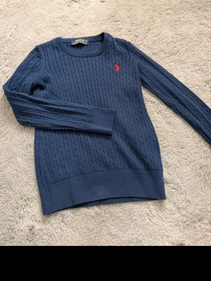 Mörkblå stickad tröja från Polo Ralph Lauren - Mörkblå stickad tröja från Polo Ralph Lauren med klassiskt kabelstickat mönster och röd broderad logotyp på bröstet. Rund halsringning och långa ärmar. Perfekt för dig som gillar stilrena och tidlösa plagg.