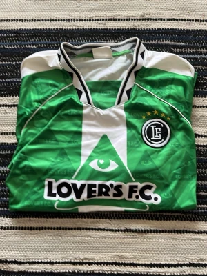 Grön Lover's F.C. fotbollströja - Snygg grön och vit fotbollströja från Lover's F.C. med svartvitt klubbmärke på bröstet och tre stjärnor ovanför. Tröjan har ett grafiskt öga på framsidan och svartvitt mönstrad krage. Perfekt för dig som vill sticka ut på planen.