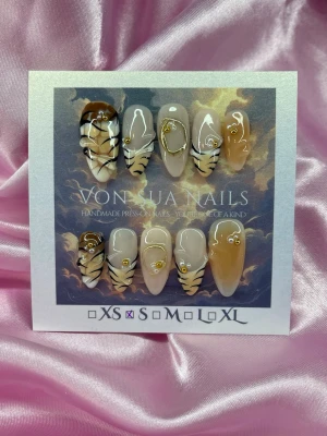 SoftClaw (S);Medim Almond - Unika handgjorda press-on naglar i tigerinspirerad design. Färgerna går i beige, brunt, svart och vitt med gulddetaljer och swirl-mönster. Formen är medium längd i almond och i storlek S: 15 mm, 11,5 mm, 12 mm, 11 mm, 9 mm . Perfekt för dig som vill sticka ut med coola detaljer.