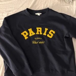 Slutsåld Populär H&M Paris tröja - Mörkblå sweatshirt med gul text 'PARIS'. Klassisk rund halsringning och ribbade muddar vid ärmslut och nederkant. Perfekt för en avslappnad och trendig look. Storlek xxs men passar även för Xs
