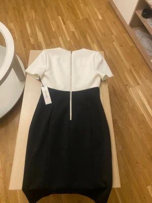 Svartvit kortklänning från Calvin Klein  - Stilren svartvit klänning från Calvin Klein med korta ärmar och markerad midja. Klänningen har en elegant tvåfärgad design, dragkedja i ryggen och är tillverkad i ett mjukt, strukturerat material. Perfekt för dig som gillar klassisk och modern stil.