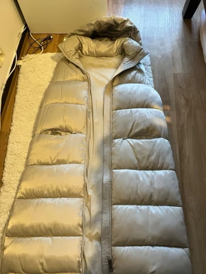 Beige lång dunväst Massimo Dutti - Lång, beige dunväst från Massimo Dutti med stor huva och quiltad design. Västen har en rak passform och dragkedja framtill. Perfekt för lager på lager och ger en clean, modern look. Materialet är glansigt och känns lyxigt.