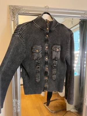 Ribbad svart kofta med denimdetaljer - Säljer en svart ribbad kofta med hög krage och knappstängning framtill. Koftan har två bröstfickor och dekorativa denimdetaljer med knappar. Långärmad och i stretchigt material som ger en cool och unik look. Väldigt stretchig, så passar många storlekar, S-L kanske.🤗