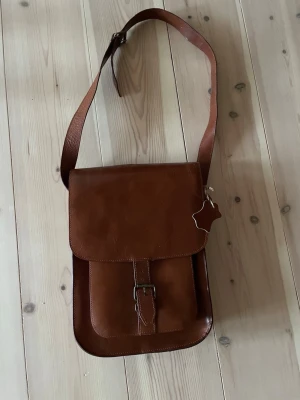 Brun axelväska i läder - Snygg brun axelväska i äkta läder med justerbar rem och klassiskt spänne framtill. Inne i väskan finns en ficka med dragkedja se bild. Väskan har en stilren, rektangulär form och en enkel design som passar till det mesta. Perfekt för dig som gillar tidlös och clean stil. Väskan kommer från Indiska.