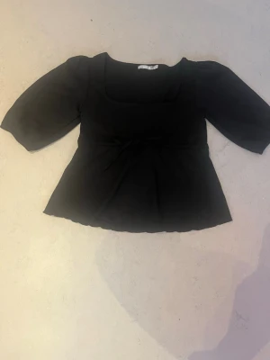 Svart topp med knyt och puffärm - Snygg svart topp från lager 157 med puffiga trekvartsärmar och en söt knytning i midjan. Toppen har en fyrkantig ringning och är lätt utsvängd nedtill, vilket ger en feminin siluett. Perfekt att styla med jeans eller kjol. Aldrig Andvänt prislappen är av 