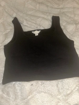 Svart ribbad linnetopp - Snygg svart ribbad linnetopp med breda axelband och fyrkantig ringning. Perfekt att styla till jeans eller kjol för en enkel och trendig look. Materialet är mjukt och stretchigt för skön passform.