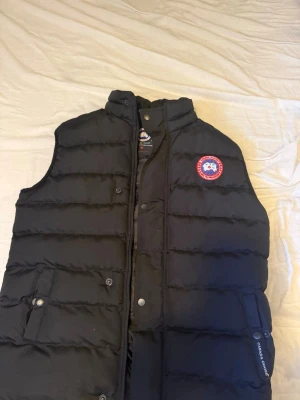 Svart dunväst från Canada Goose - Snygg svart dunväst från Canada Goose med klassisk logotyp på bröstet. Västen har ståkrage, dragkedja och knappar framtill samt tre fickor varav en på insidan. Storlek S men kan passa M med. Jag är även öppen för att diskutera pris ifall det önskas.