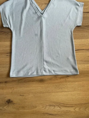 Ljusgrå randig v-ringad topp - En ljusgrå topp med tunna diagonala ränder och v-ringning. Modellen är kortärmad och har en rak, loose passform. Tyget känns mjukt och lätt, perfekt för en avslappnad stil. En fläck som syns i bilden
