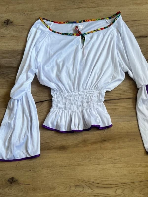 Vit cinched  blus med färgglada detaljer - Säljer en vit blus från Costume med långa puffiga ärmar och smockad midja. Halsringningen har färgglada mönstrade kanter och en liten knytning framtill. Lila kant längst ner på ärmar och nederkant. Perfekt för en trendig och lekfull look. Ny skick