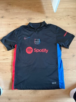  FC Barcelona fotbollströja borta Raphinha 24/25 - Snygg svart FC Barcelona matchtröja med röda och blå detaljer på sidorna. Tröjan har Spotify-logga på bröstet, klubbmärke och Nike-logga. På ryggen står det Raphinha och nummer 11 i rött. Tillverkad i lätt och ventilerande material, perfekt för fotboll. Pris kan diskuteras. 