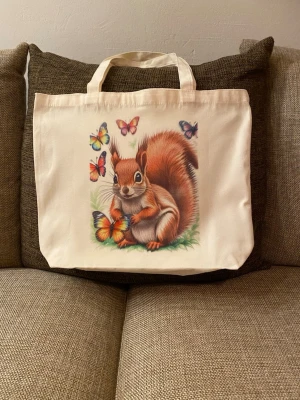 Tygkasse/shoppingväska med ekorre och fjärilar - Supersöt shoppingväska i cremefärgad tyg med tryck av en ekorre omgiven av färgglada fjärilar. Väskan är perfekt för shopping, för att bära böcker, mat eller annat smått och gott. 