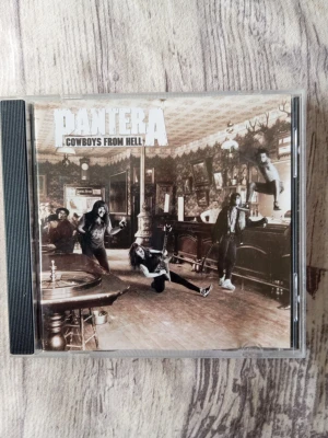 Pantera Cowboys From Hell CD - CD-albumet 'Cowboys From Hell' av Pantera i original plastfodral. Omslaget har ett sepiafärgat foto med bandmedlemmarna i en bar. Själva CD:n är ljusrosa med gul text och låtlista tryckt på framsidan. Perfekt för dig som gillar metal och vill ha klassisk musik på fysisk skiva.