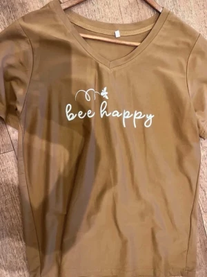 En NY Brun v-ringad t-shirt Bee Happy - Snygg brun t-shirt med v-ringning och vit texten 'bee happy' framtill tillsammans med en liten bi-detalj. T-shirten har korta ärmar och är gjord i mjuk bomull, perfekt för en chill och avslappnad stil.