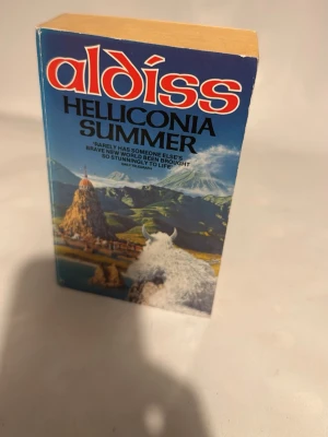 Helliconia Summer av Brian Aldiss - Science fiction-roman med färgstarkt omslag i blått och rött. Boken är en pocket med illustration av berg, en vit yxa och en fantasifull värld på framsidan. Perfekt för dig som gillar episka äventyr och framtidsvisioner.