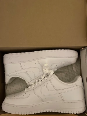 Nike Air Force 1 vita sneakers - Klassiska helvita Nike Air Force 1 sneakers i lågt utförande. Skorna har en stilren design med perforerad tå, vita skosnören och den ikoniska swoosh-loggan på sidan. Tillverkade i läder med gummisula för bra komfort och streetkänsla.