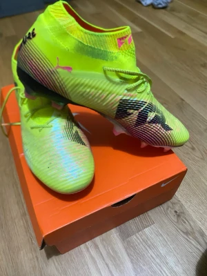 Neongula Puma Future fotbollsskor - Säljer ett par neongula Puma Future ultimate fotbollsskor med svart och rosa grafik. Skorna har stickad strumpkrage för extra stöd, snörning och en lätt syntetisk ovandel. Perfekta för dig som vill sticka ut på planen och ha bra grepp i varje steg.