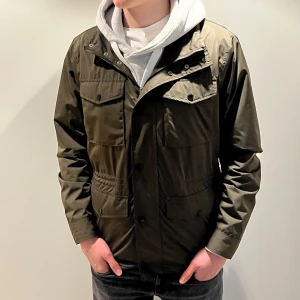 Mörkgrön vindjacka med fickor - Stilren mörkgrön fieldjacket med fyra framfickor och knappdetaljer. Jackan har hög krage och dragkedja framtill, samt en rak passform som passar till många olika looks. Perfekt för dig som vill ha en enkel men snygg jacka till vardags.