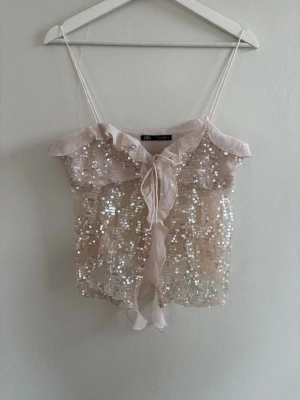 Paljettlinnetopp från Zara med volang - Supersöt ljusbeige linnetopp från Zara med tunna axelband, volangkant och knyt framtill. Toppen är täckt av glittriga paljetter som ger ett extra lyft till outfiten. Perfekt för dig som vill sticka ut lite extra.