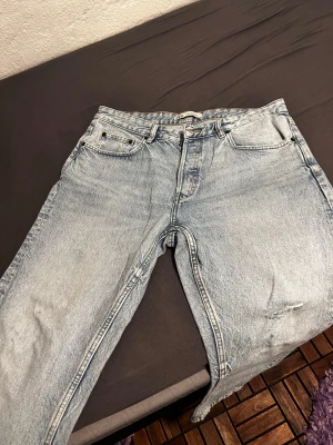 Ljusblå raka jeans med slitningar - Säljer ett par ljusblå jeans med raka ben och snygga slitningar på låret. Jeansen har klassisk femficksdesign och dragkedjegylf. Perfekta för en avslappnad och trendig look.