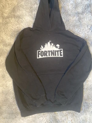 Svart Fortnite hoodie med tryck - Säljer en svart hoodie med stor vit Fortnite-logga och siluett på bröstet. Klassisk känguruficka framtill och huva. Perfekt för dig som gillar gaming och vill ha en chill och snygg stil. Mjuk insida och ribbade muddar vid ärmar och nederkant.