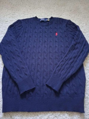Mörkblå kabelstickad tröja Polo Ralph Lauren - Kabelstickad mörkblå tröja från Polo Ralph Lauren med klassisk rund halsringning och röd broderad logga på bröstet. Tröjan har ribbade muddar vid ärmslut och nederkant. Perfekt för dig som gillar tidlös och stilren design. Passar lmer som S.