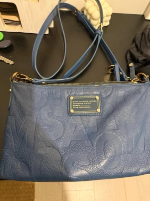 Blå axelväska Marc by Marc Jacobs - Snygg blå axelväska från Marc by Marc Jacobs i präglat läder med stora bokstäver och guldfärgade detaljer. Väskan har justerbar axelrem, dragkedja upptill och en extra ficka med dragkedja på baksidan. Perfekt för dig som gillar stilrena accessoarer.