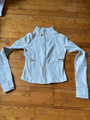 Vit sportig jacka från Lululemon - Snygg vit jacka från Lululemon med hög krage och dragkedja framtill. Jackan har två fickor med dragkedja och en figurnära passform. Tillverkad i stretchigt syntetmaterial, perfekt för en aktiv vardag.