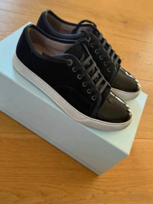  Lanvin Captoe  - Snygga sneakers från Lanvin i svart och mörkblå mocka med lackade tåpartier och vita sulor. Skorna har klassisk låg profil, matchande skosnören och metallöljetter. Perfekta för dig som vill ha en stilren och exklusiv look. Storlek 43