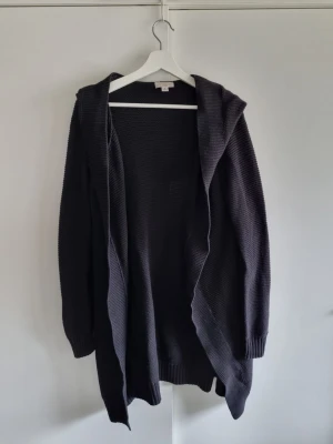 Svart lång kofta med huva Witchery M - Mysig svart lång kofta från Witchery med huva och öppen front. Stickad i ett grovt mönster som ger en avslappnad look. Perfekt att slänga över en t-shirt eller topp för extra lager. Ribbstickade muddar vid ärmslut och nederkant. 100% bomull. Knappt använd. 