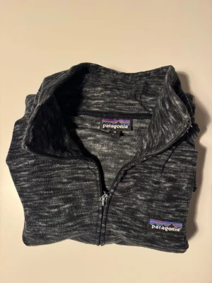 Patagonia full-zip fleece svart melange, M, herr - Patagonia full-zip i mjuk mikrofleece, svart/grå melange. Ståkrage, två handfickor. Gott skick. litet hål på ärmen (myrstort, se bild 5). Stl M herr.  Skicka mig gärna era erbjudanden