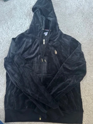 Svart velour hoodie från U.S. Polo Assn - Mjuk och skön svart hoodie i velour från U.S. Polo Assn med dragkedja framtill, huva med snören och guldfärgad logga på bröstet. Stora fickor framtill och ribbade muddar vid ärmslut och nederkant. Perfekt för en chill och avslappnad stil.
