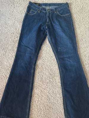 Blå bootcut jeans från Lee - Säljer ett par klassiska blå bootcut jeans från Lee med fem fickor och kontrastsömmar. Jeansen har normal midja och är tillverkade i slitstark denim. Perfekta för dig som gillar retrostil och vill ha ett par tidlösa jeans.