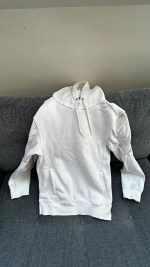 Vit hoodie från Club de Demain - Säljer en stilren vit hoodie från Club de Demain med broderad logga på bröstet. Hoodien har dragsko i huvan och en klassisk känguruficka framtill. Tillverkad i mjukt bomullsmaterial som är perfekt för chill dagar eller när du vill ha en clean look.