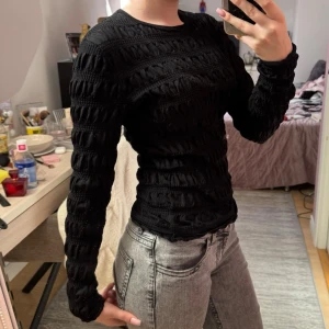 Svart ribbad långärmad tröja - Säljer en svart långärmad tröja med ribbad struktur och diskreta vågiga detaljer. Tröjan har en kroppsnära passform och rund halsringning. Perfekt för dig som gillar stilrena och trendiga plagg.