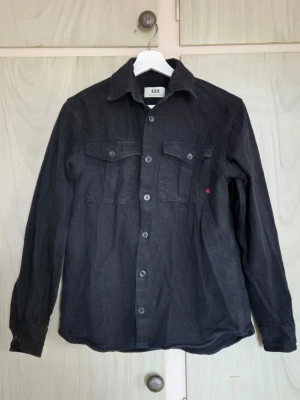 Svart denim skjorta från RDD med fickor - Svart denim skjorta från Royal Denim Division med två bröstfickor med knappar och en liten röd detalj på ena fickan. Skjortan har klassisk krage, långa ärmar och är tillverkad i ett denim. Perfekt att bära öppen eller stängd.  Otroligt bra skick använd 1-2 gånger. Extraknapparna finns kvar.                           Alla kläder rollas innan dom skickas hem då det finns katt i hemmet🐈 Jag stryker och tvättar INTE innan jag skickar det får du stå för själv så det blir som du vill ha det🤗