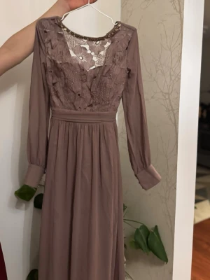 Lång brun klänning med spetsdetaljer - Romantisk långklänning i brun ton med långa ärmar och vacker spets upptill. Klänningen har volangdetaljer på bröstet och en markerad midja. Perfekt för dig som gillar en bohemisk och elegant stil.