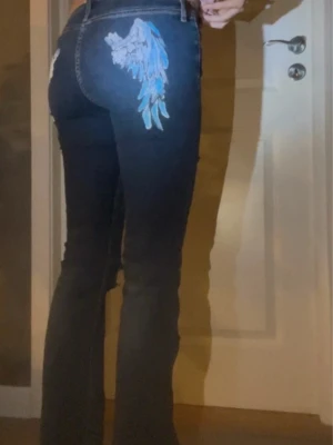 Mörkblå jeans med blå änglavingar - Säljer ett par mörkblå jeans med unik broderad änglavinge i blått och vitt på bakfickan. Jeansen har bootcut-modell och klassisk femficksdesign. Snygga kontrastsömmar och slitna detaljer vid benslut. Perfekta för dig som vill sticka ut med coola detaljer. Unik design som är custom made.