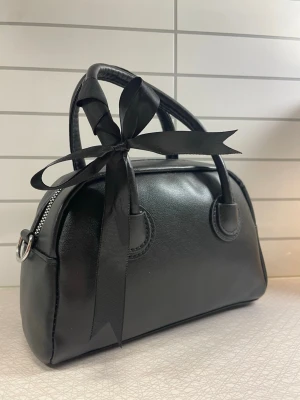 Svart handväska med rosett - Stilren svart handväska (crossbody) i fejkläder med rundad form och dubbla handtag. Väskan har en dekorativ svart rosett på ena handtaget.    Aldrig använd.                                                   Leverans med Instabox eller mötesplats. 