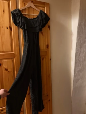 Svart glittrig off shoulder jumpsuit - Snygg svart jumpsuit med glittereffekt och off shoulder-krage med volang. Vida ben och markerad midja ger en trendig siluett. Perfekt för dig som vill sticka ut lite extra. Materialet känns mjukt och följsamt.