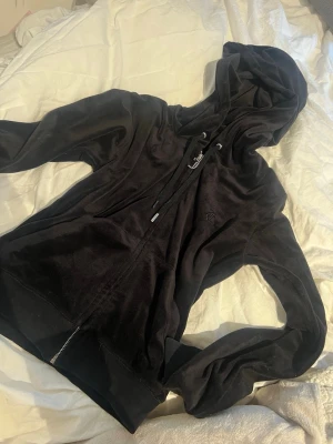 Svart velour juicy couture tröja - Mjuk och skön svart hoodie i velour med dragkedja framtill och justerbar huva med snören. Två fickor på sidorna och diskret broderad logga på bröstet. Perfekt för en chill och avslappnad stil.