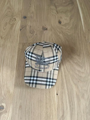 Rutig Burberry keps i beige och svart - Säljer en klassisk rutig keps från Burberry i beige, svart och vitt. Kepsen har det ikoniska Burberry-mönstret och broderad logga framtill. Perfekt accessoar för dig som vill ha en tidlös och trendig look.