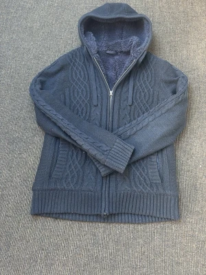 Mörkblå stickad hoodie med dragkedja - Mysig mörkblå hoodie med stickat flätmönster och dragkedja framtill. Insidan är fodrad med mjuk teddy för extra värme. Huva med dragsko och ribbade muddar vid ärmslut och nederkant. Perfekt för kalla dagar och riktigt skön att ha på sig. Jätte fint skick storlek L