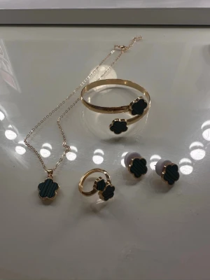 Set med fyrklöver smycken i guld - Snyggt smyckeset med halsband, armband, ring och örhängen i guldfärg. Alla delar har mörkgröna fyrklöverformade hängen med räfflad yta. Perfekt för dig som gillar unika accessoarer med elegant touch.