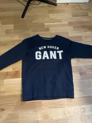 Mörkblå sweatshirt från GANT - Mörkblå sweatshirt från GANT med trycket 'NEW HAVEN GANT' i vitt på bröstet. Klassisk rund halsringning och långa ärmar. Perfekt för en avslappnad och stilren look.