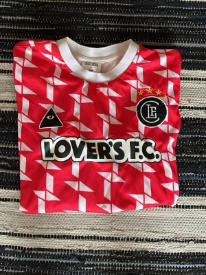 Röd Lover's F.C. fotbollströja - Snygg röd fotbollströja från Lover's F.C. med vitt geometriskt mönster, svart och vitt tryck på bröstet samt klubbmärke med tre stjärnor. Tröjan har vit rund halsringning och är tillverkad i ett lätt, sportigt material.