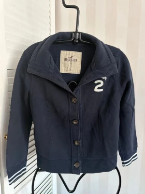 Mörkblå kofta från Hollister - Mörkblå kofta från Hollister med knappar framtill och vit broderad logga samt siffran 2 på bröstet. Tröjan har ribbade muddar med vita ränder och en hög krage som kan vikas ner. Perfekt för en avslappnad och sportig stil.