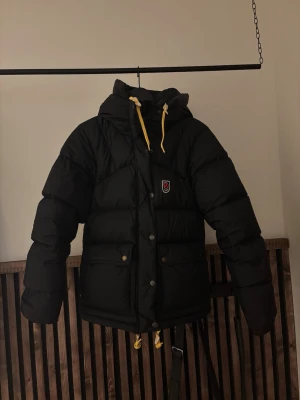 Svart dunjacka från Fjällräven - Säljer en svart dunjacka från Fjällräven med huva och gula dragskor. Jackan har två stora fickor framtill, tryckknappar och dragkedja samt Fjällrävens klassiska logga på bröstet. Perfekt för kalla vinterdagar.