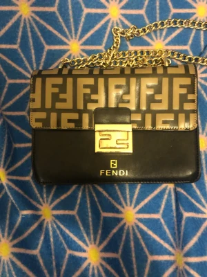 Svart och beige Fendi handväska - Säljer en snygg handväska från Fendi med klassiskt FF-mönster i beige och svart. Väskan har guldfärgad kedja som axelrem och ett stilrent spänne framtill. Materialet är skinn och insidan har en praktisk dragkedja. Perfekt för dig som gillar lyxiga detaljer och ikonisk design.